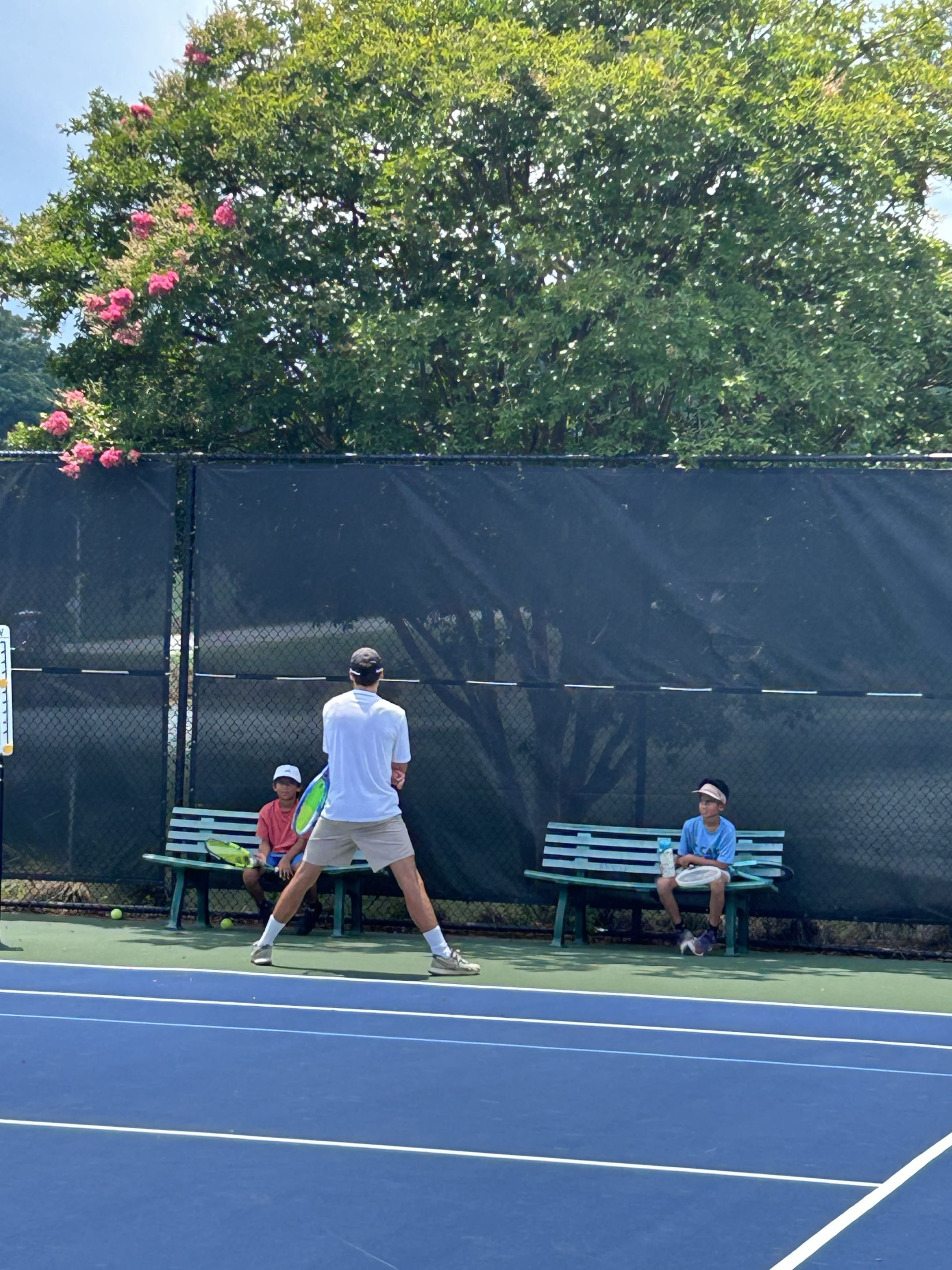 Coaching session on an outdoor tennis court with two junior players watching a demonstration.